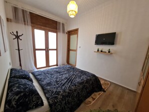 A bedroom with a bed, a TV mounted on the wall, a window with curtains, and a wooden door. - Anir House (Tanger)