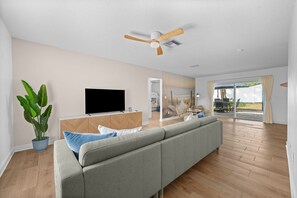 Ein geräumiger Wohnbereich mit Flachbildfernseher, einem grauen Sofa, Deckenventilator und einer großen Topfpflanze.