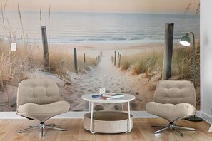 Ein Zimmer mit einem Strandgemälde, zwei Stühlen, einem kleinen runden Tisch und einer Lampe.