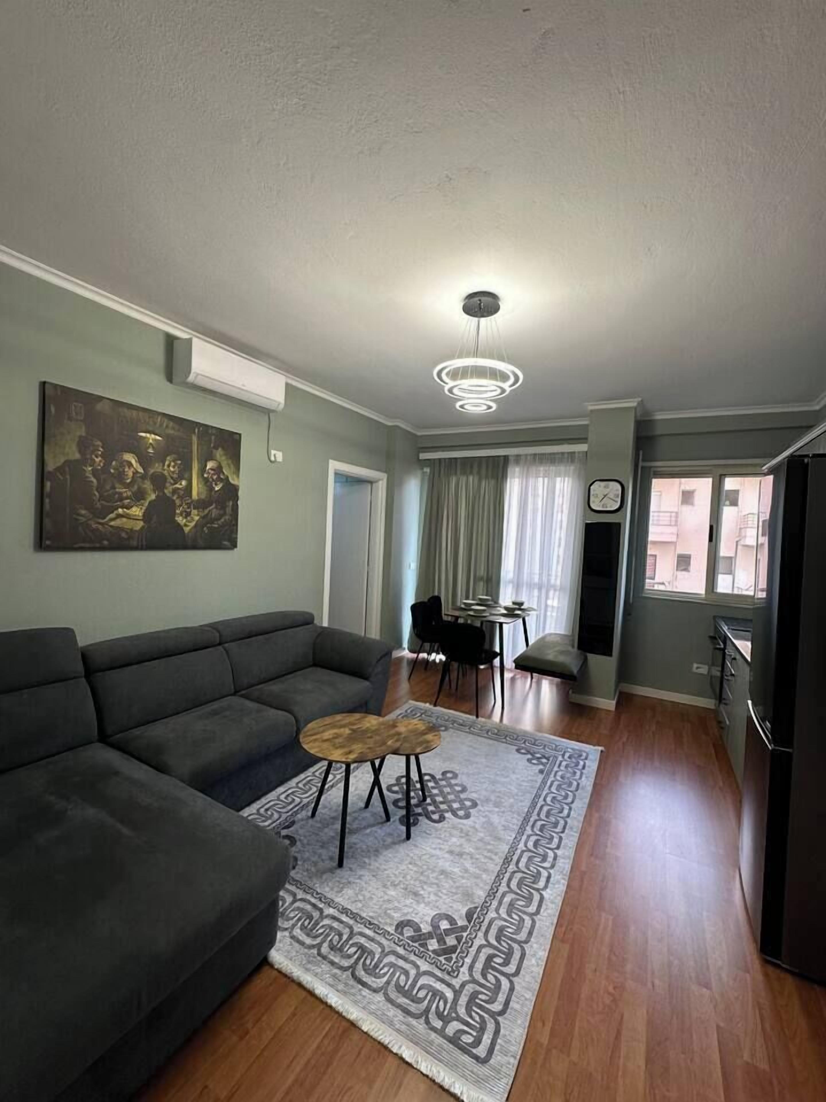 Een woonkamer met een bank, een salontafel en een vloerkleed. Er is een eethoek met stoelen en een tafel. Een klok hangt aan de muur.