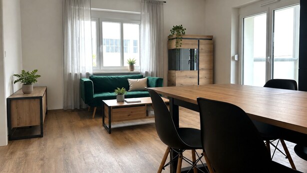 A modern, well-lit room with a green sofa, wooden dining table, and chairs. There is a small side table with a plant, and a cabinet with a mirror. - Stylish dream apartment - 2 bedrooms - balcony (Bayreuth)