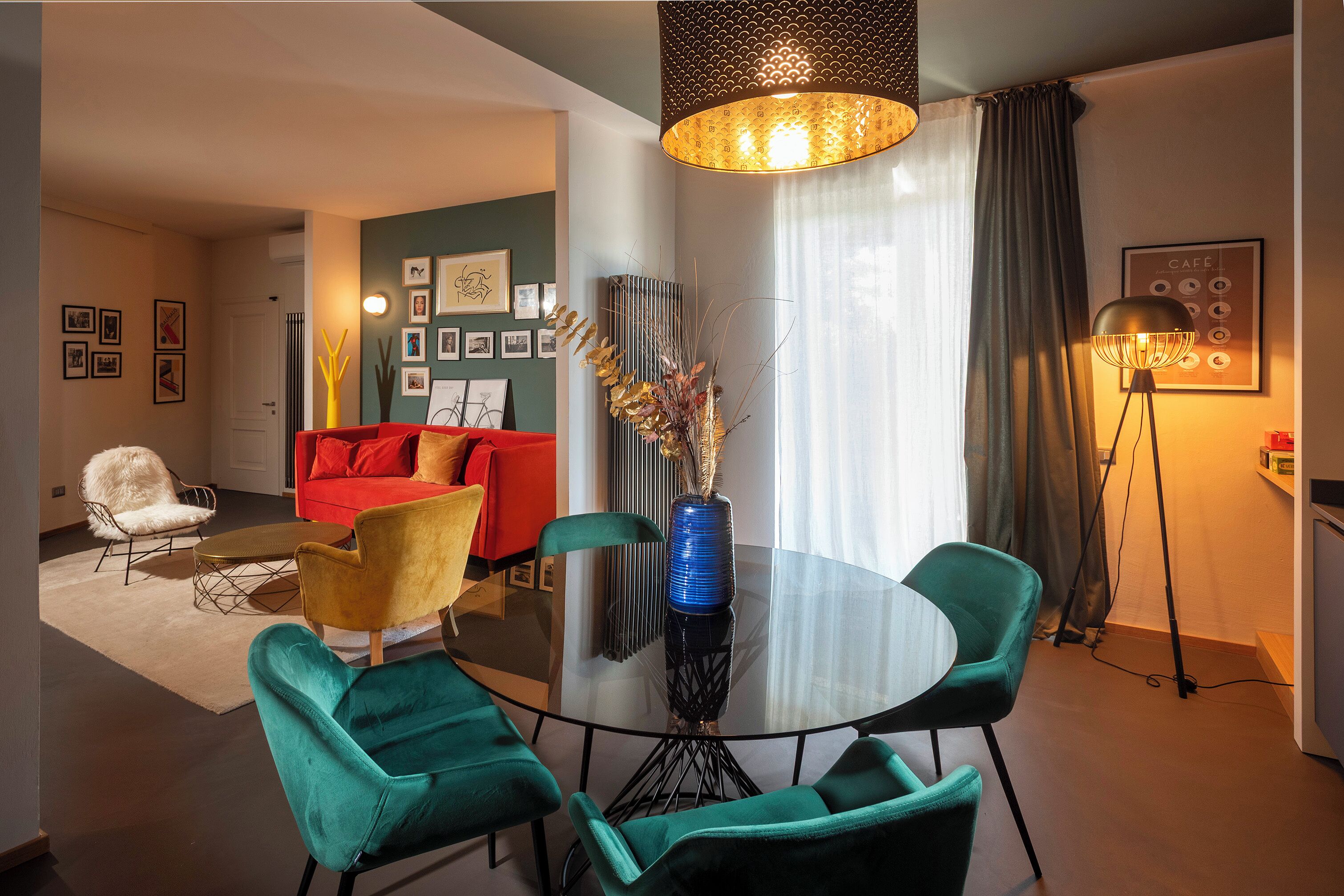 A modern living room with a round glass table, teal chairs, a red sofa, and a large vase with decorative branches.