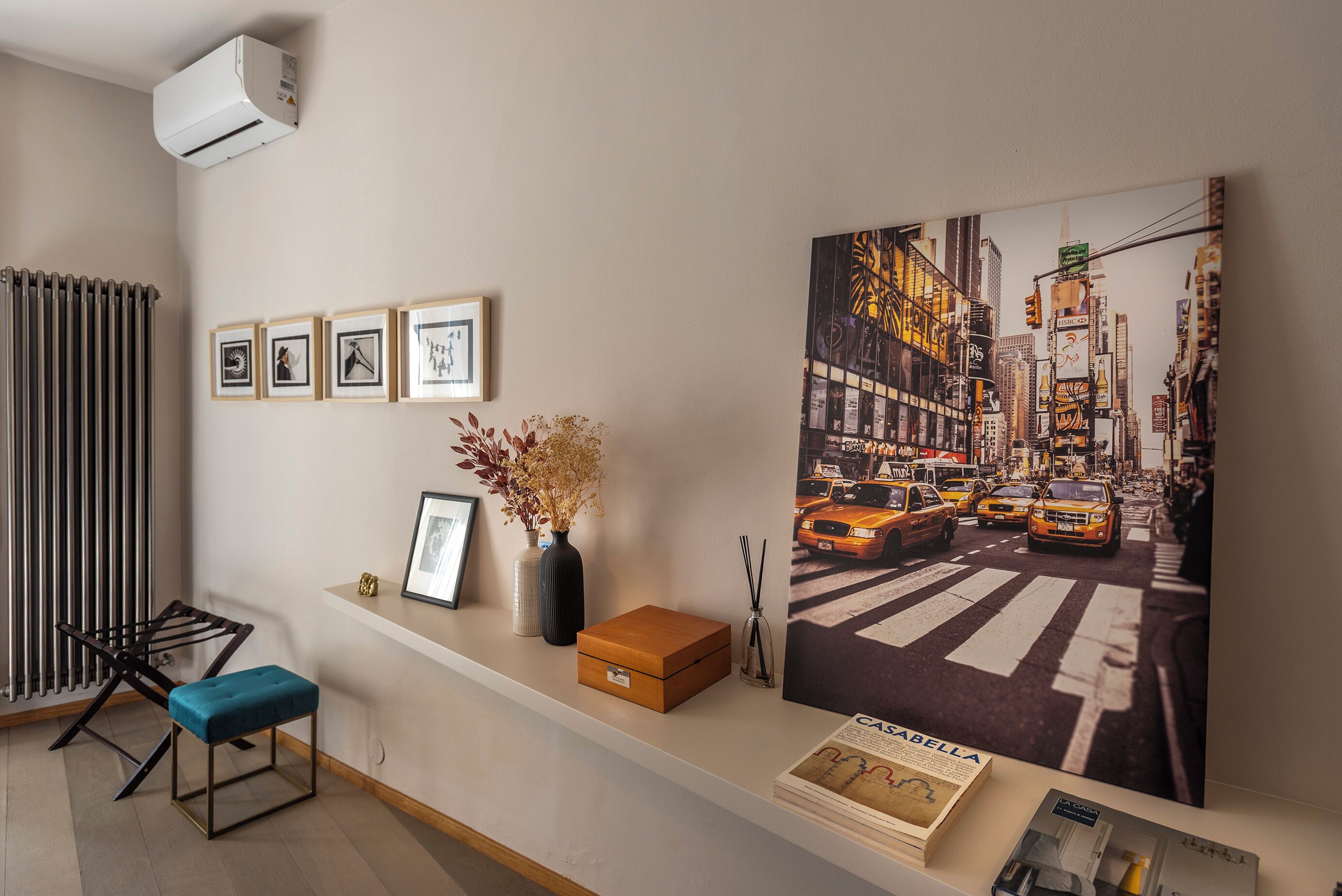 Een kamer met een radiator, ingelijste foto's, een vaas met gedroogde bloemen, een bankje, een doos en een tijdschrift op een plank.