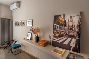 Ein Zimmer mit Heizkörper, gerahmten Bildern, einer Vase mit Trockenblumen, einer Bank, einer Kiste und einer Zeitschrift auf einem Regal.