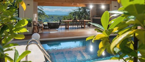 A pool area with a wooden deck, a table with chairs, and a view of the landscape.