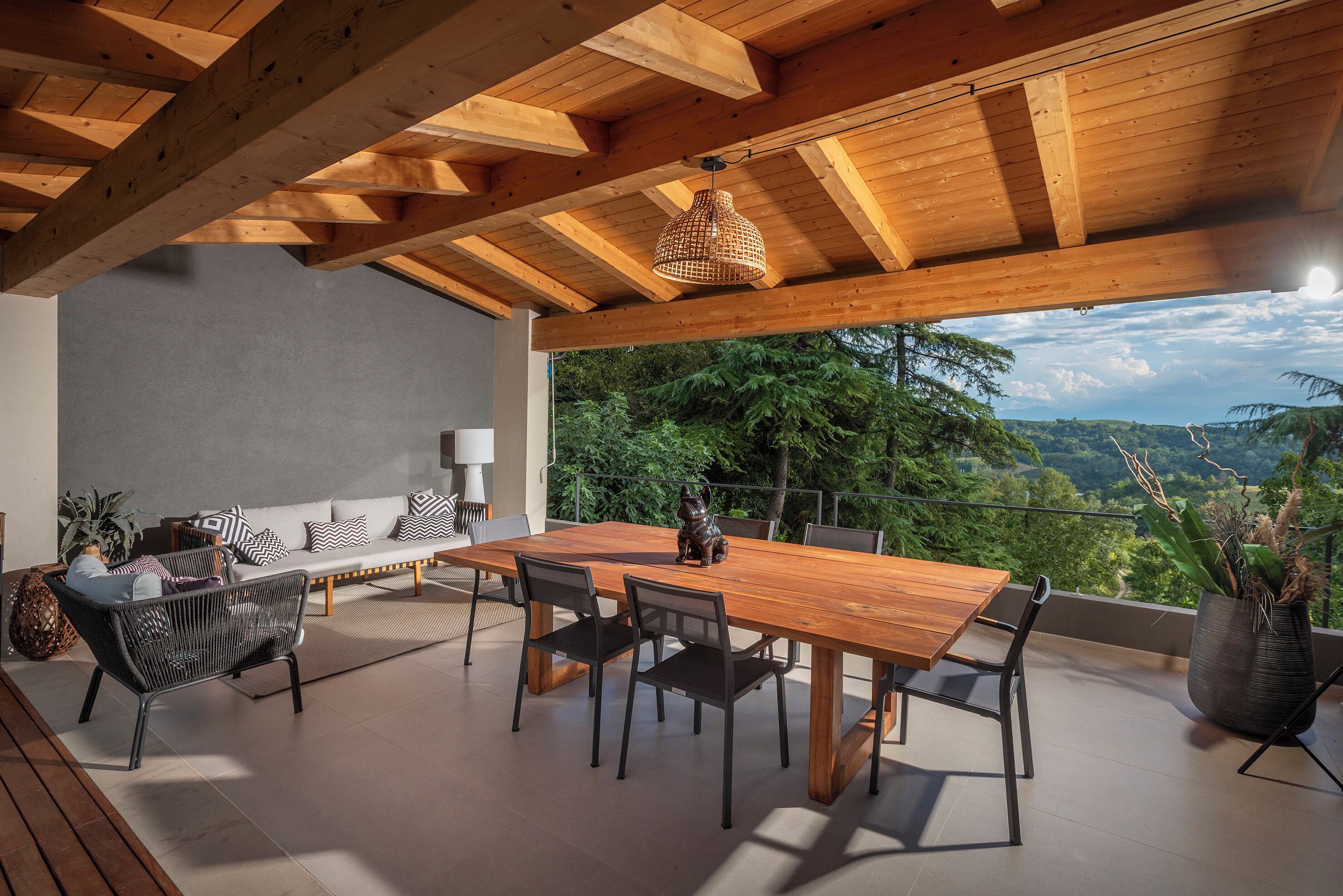 A wooden dining table with chairs, a sofa, and a hanging light fixture in a covered outdoor area with a view of trees and hills.