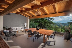 Mesa de madera con sillas, un sofá y una lámpara colgante en un espacio exterior cubierto con vistas a árboles y colinas.