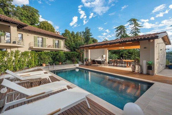 Una piscina con sillones de descanso, una terraza de madera y un área de comedor al aire libre techada.