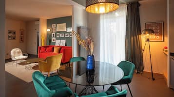 A modern living room with a round glass table, teal chairs, a red sofa, and a large vase with decorative branches.