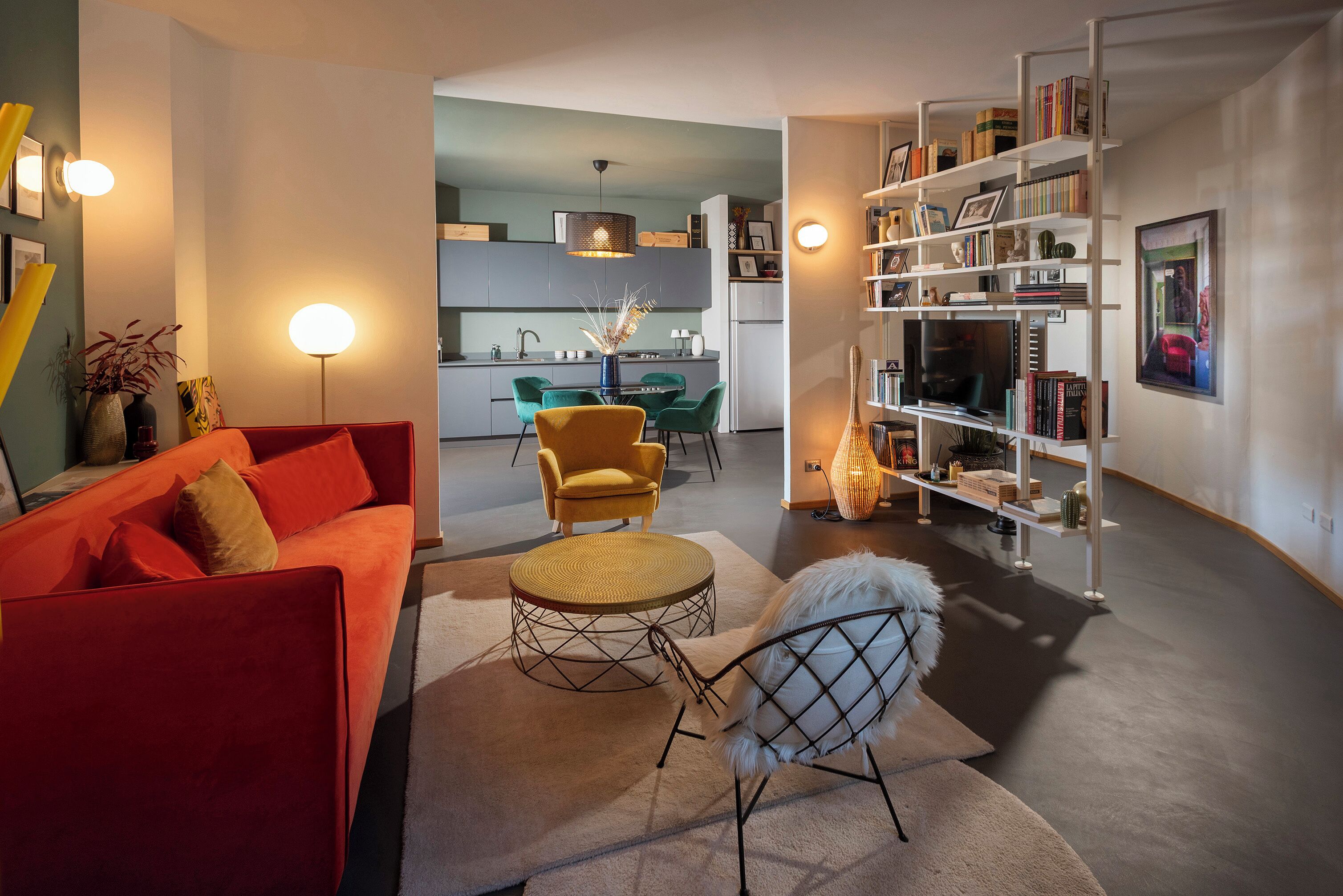A modern living room with a red sofa, a yellow armchair, and a white chair with a fur throw. A bookshelf filled with books and decorative items is visible.