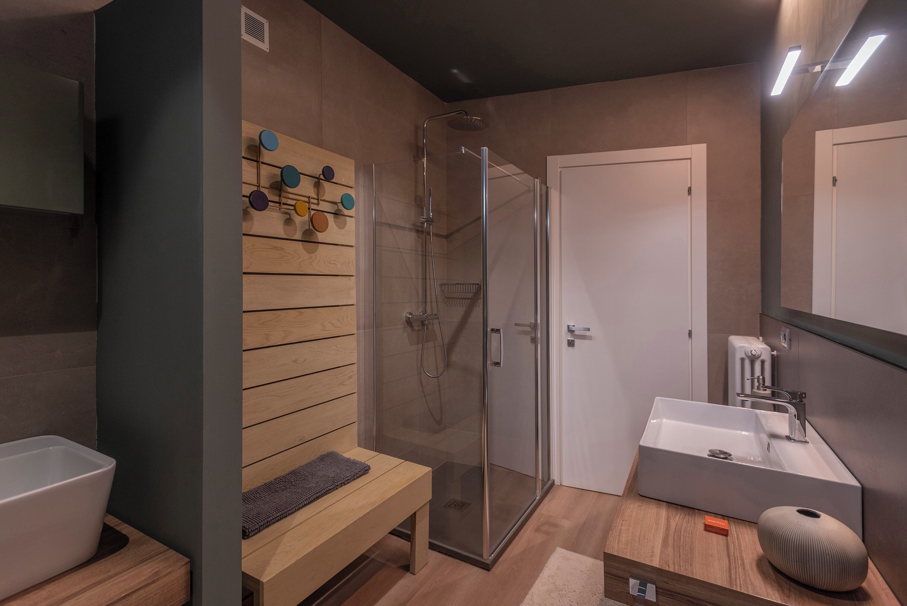 A modern bathroom with a glass-enclosed shower, a wooden bench, a sink, and a mirror.