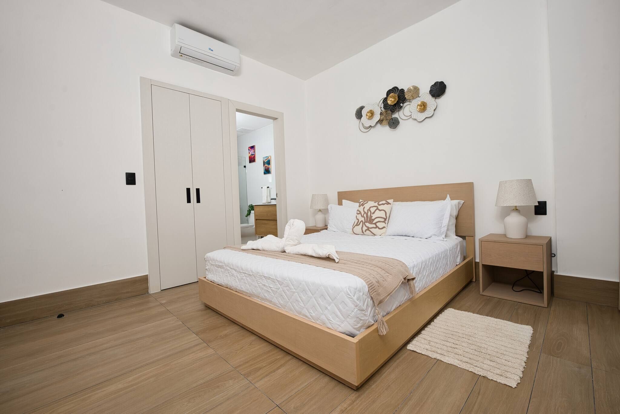 A modern bedroom with a wooden bed, white bedding, a bedside table with a lamp, and a wall-mounted air conditioner.