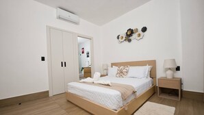 A modern bedroom with a wooden bed, white bedding, a bedside table with a lamp, and a wall-mounted air conditioner. - Modern Apartment With Private Pool in Punta Cana (Punta Cana)