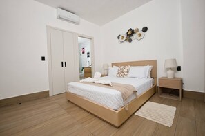 Ein modernes Schlafzimmer mit einem hölzernen Bett, weißer Bettwäsche, einem Nachttisch mit Lampe und einer wandmontierten Klimaanlage.