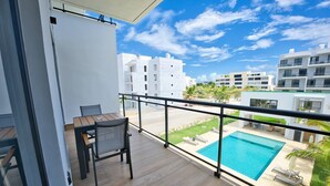 A balcony with a wooden table and chairs, overlooking a swimming pool and a landscaped area with palm trees. - Modern Apartment With Private Pool in Punta Cana (Punta Cana)