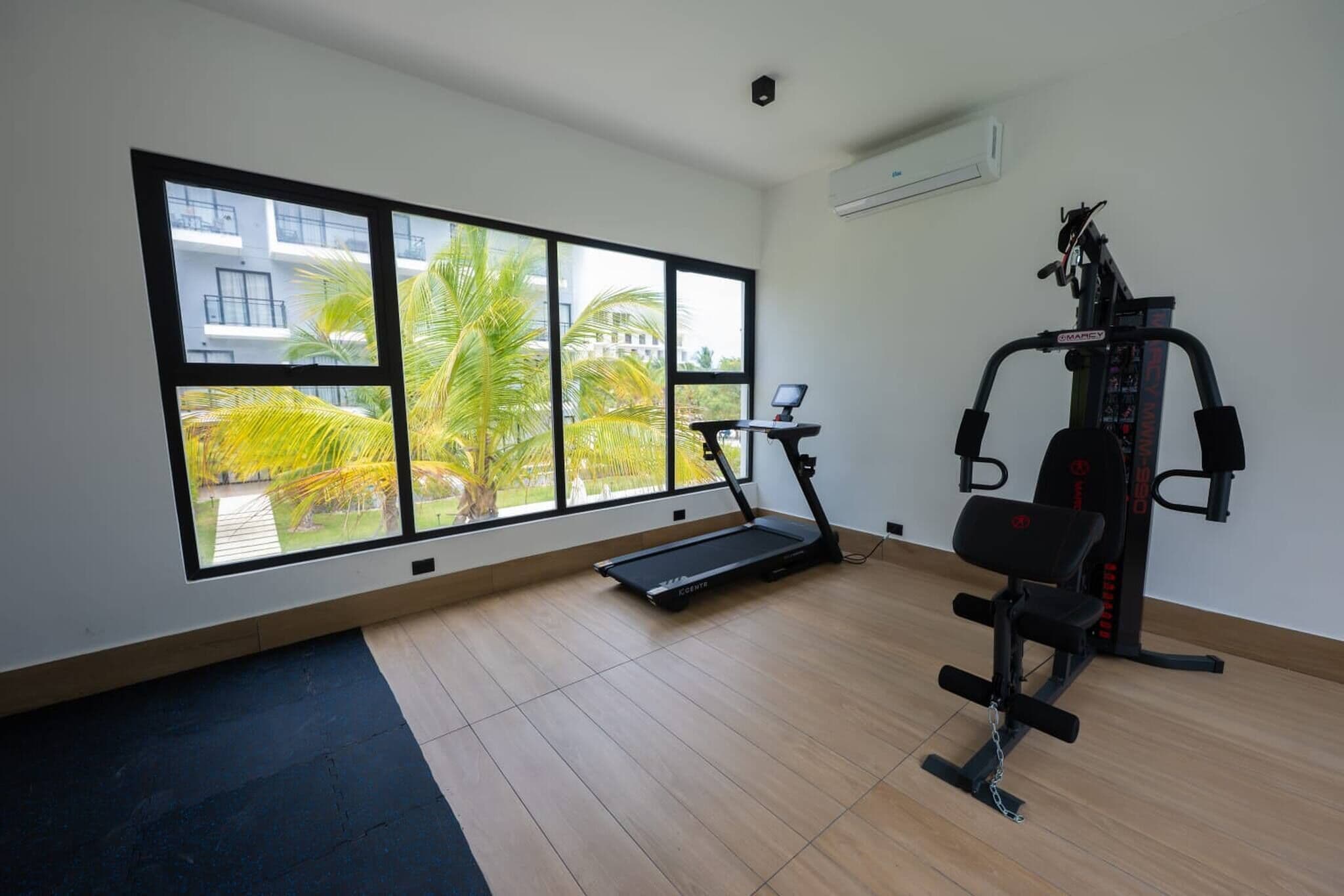 A gym with a treadmill, weight machines, and large windows overlooking a courtyard with palm trees.