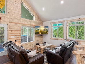Ein gemütliches Blockhauszimmer mit Holzvertäfelung, einem Flachbildfernseher, einer Couch, einem Tisch und großen Fenstern mit Blick auf Grünflächen.