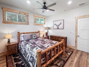 Quarto com estrutura de cama de madeira, colcha com estampas de animais, duas mesas de cabeceira com luminárias, um ventilador de teto e uma janela com persianas.
