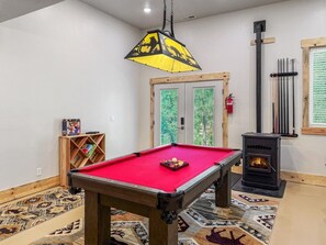 Ein Billardtisch mit roter Spielbahn, ein Holzregal mit Büchern, ein Kamin und ein Fenster mit Blick auf Bäume.