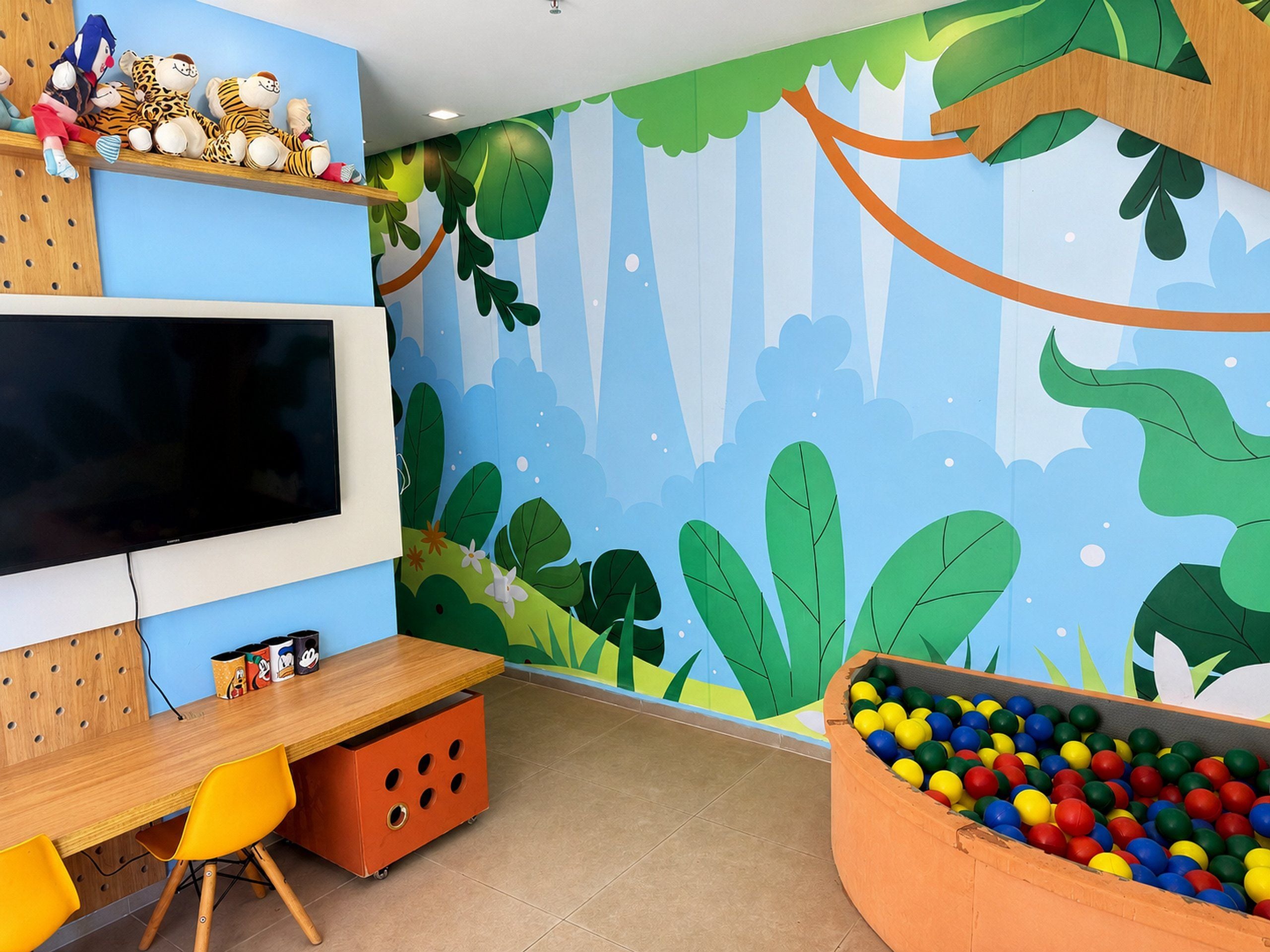 Habitación con un televisor de pantalla plana grande montado en la pared, un escritorio de madera con una computadora, un colorido arenero y un mural en la pared con una escena de bosque.