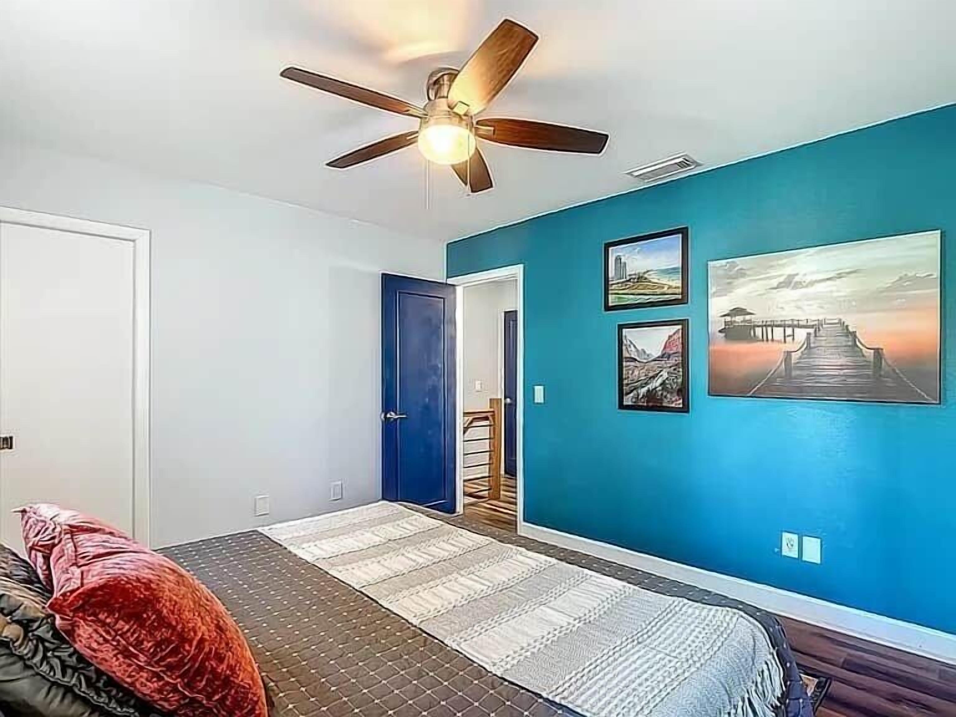 Quarto com cama, ventilador de teto, parede azul e porta de entrada para outro cômodo.