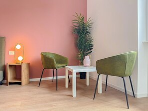A room with a pink wall, a white table, two green chairs, a mirror with lights, a potted plant, and a small wooden shelf. - Flatsharing - Gioia Navy suite - 1 bedroom (Milan)