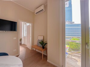A room with a flat-screen TV mounted on the wall, a wooden bench with a plant, and a view of a cityscape through a glass door. - Flatsharing - Gioia Navy suite - 1 bedroom (Milan)