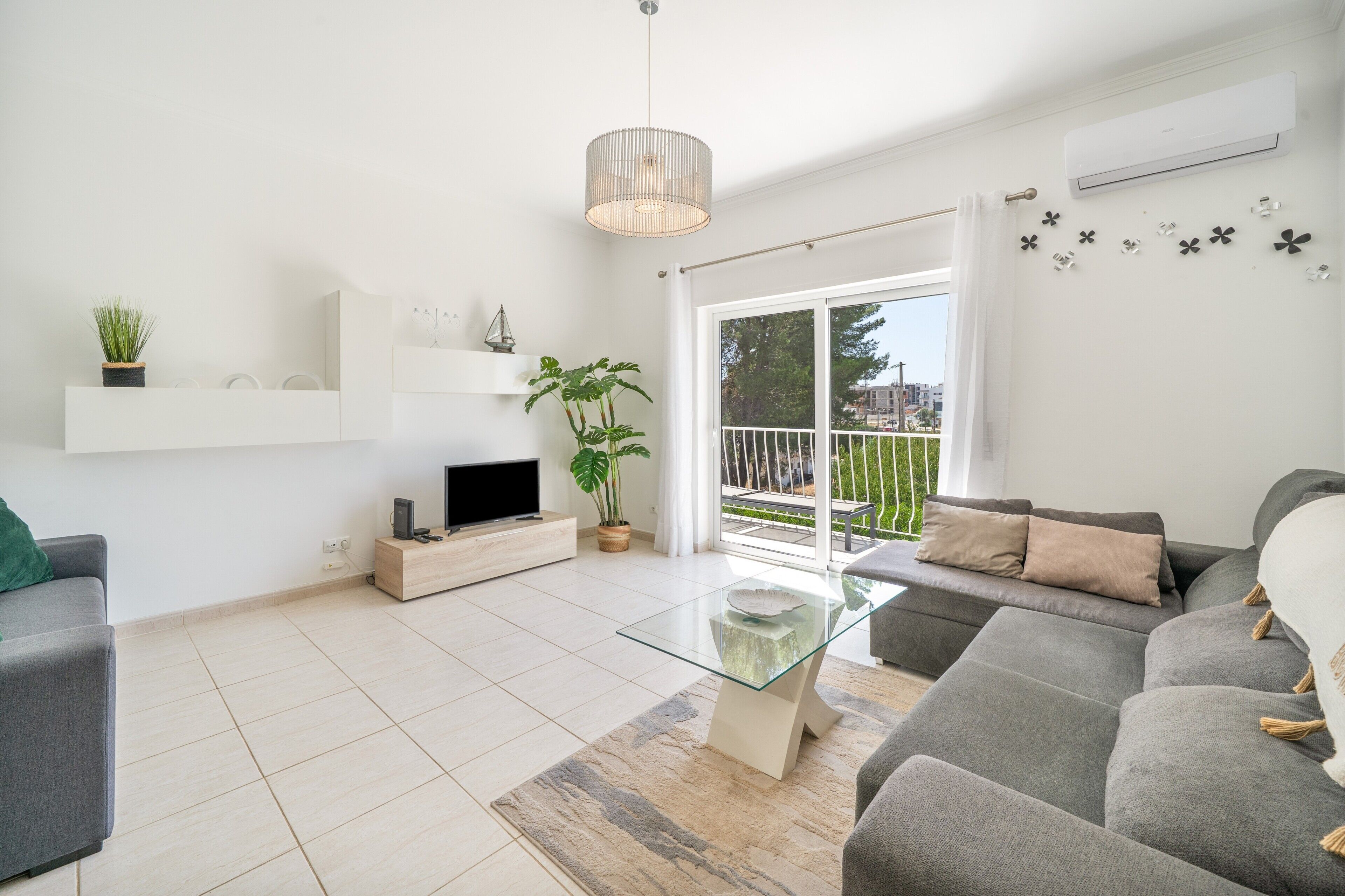 A modern living room with a grey sofa, a glass coffee table, a TV on a wooden stand, a potted plant, and a balcony with a railing.