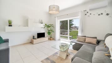 A modern living room with a grey sofa, a glass coffee table, a TV on a wooden stand, a potted plant, and a balcony with a railing.