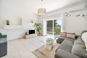 Una sala moderna con un sofá gris, una mesa de centro de vidrio, un televisor sobre un mueble de madera, una planta en maceta y un balcón con barandilla.