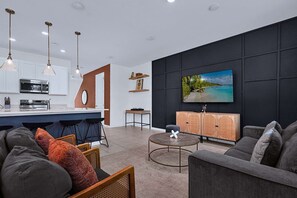 A modern living room with a large flat-screen TV, a dark accent wall, and a kitchen area with white cabinets and a bar. - Disney Home w Lake View BBQ Patio & Resort Access (Davenport)