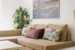 Ein beiges Sofa mit gemusterten Kissen, einer Blumentopf-Pflanze und einem gerahmten Bild einer Brücke.