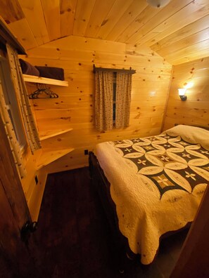 Una habitación de cabaña de madera con una cama, una lámpara colgante y una ventana con cortinas.