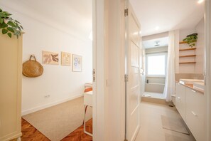 Ein Flur, der zu einem Badezimmer mit Dusche, Toilette und Waschbecken führt. An der Wand befinden sich Bilder in Rahmen, und links steht eine Pflanze.