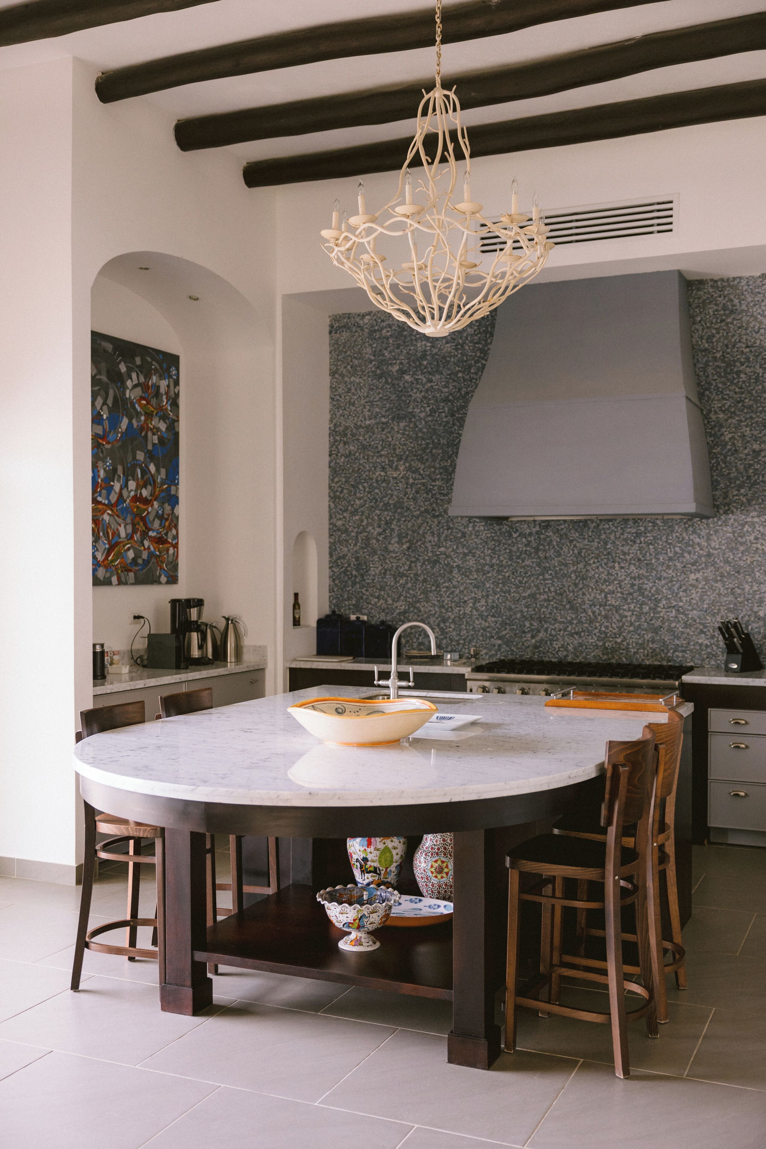 A modern kitchen with a central island, dark wooden chairs, a hanging light fixture, and a granite backsplash.