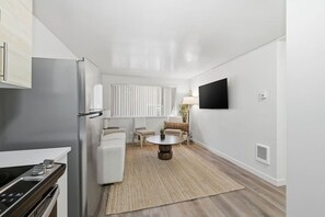 A compact kitchen with a refrigerator, stove, and microwave. There is a dining area with a table and chairs, a TV mounted on the wall, and a window with blinds.