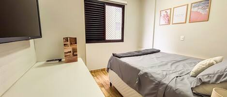 A bedroom with a bed, a desk, a window with blinds, and a wall with framed pictures.