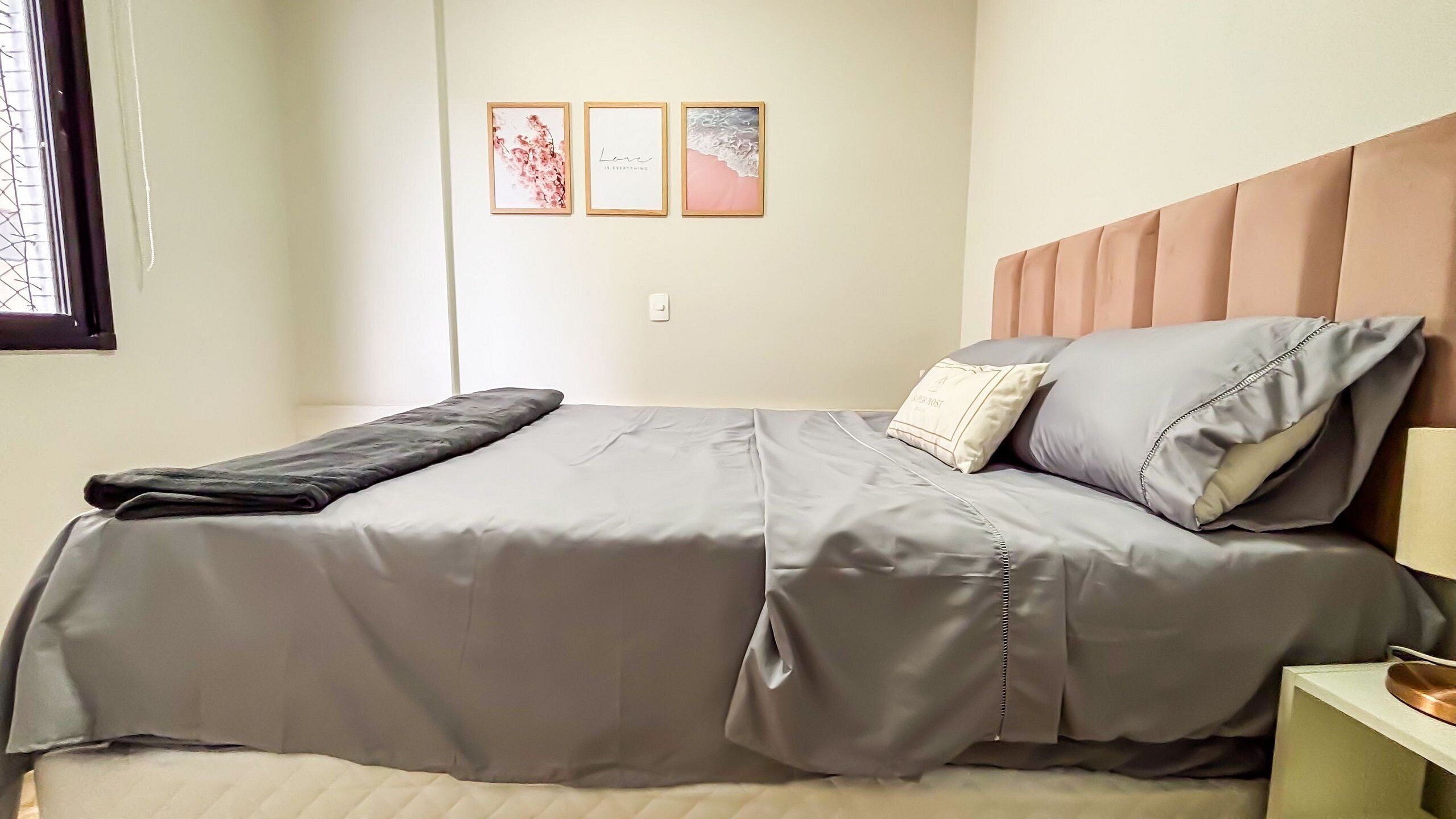 Una cama con una colcha gris, una almohada con una etiqueta y una mesita de noche con una lámpara.