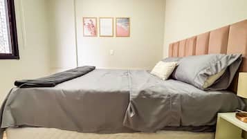Una cama con una colcha gris, una almohada con una etiqueta y una mesita de noche con una lámpara.