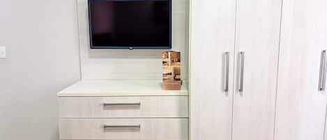 Una habitación con un televisor de pantalla plana montado en una pared de tonos claros, una pequeña caja de madera y un mueble con dos cajones y dos puertas.