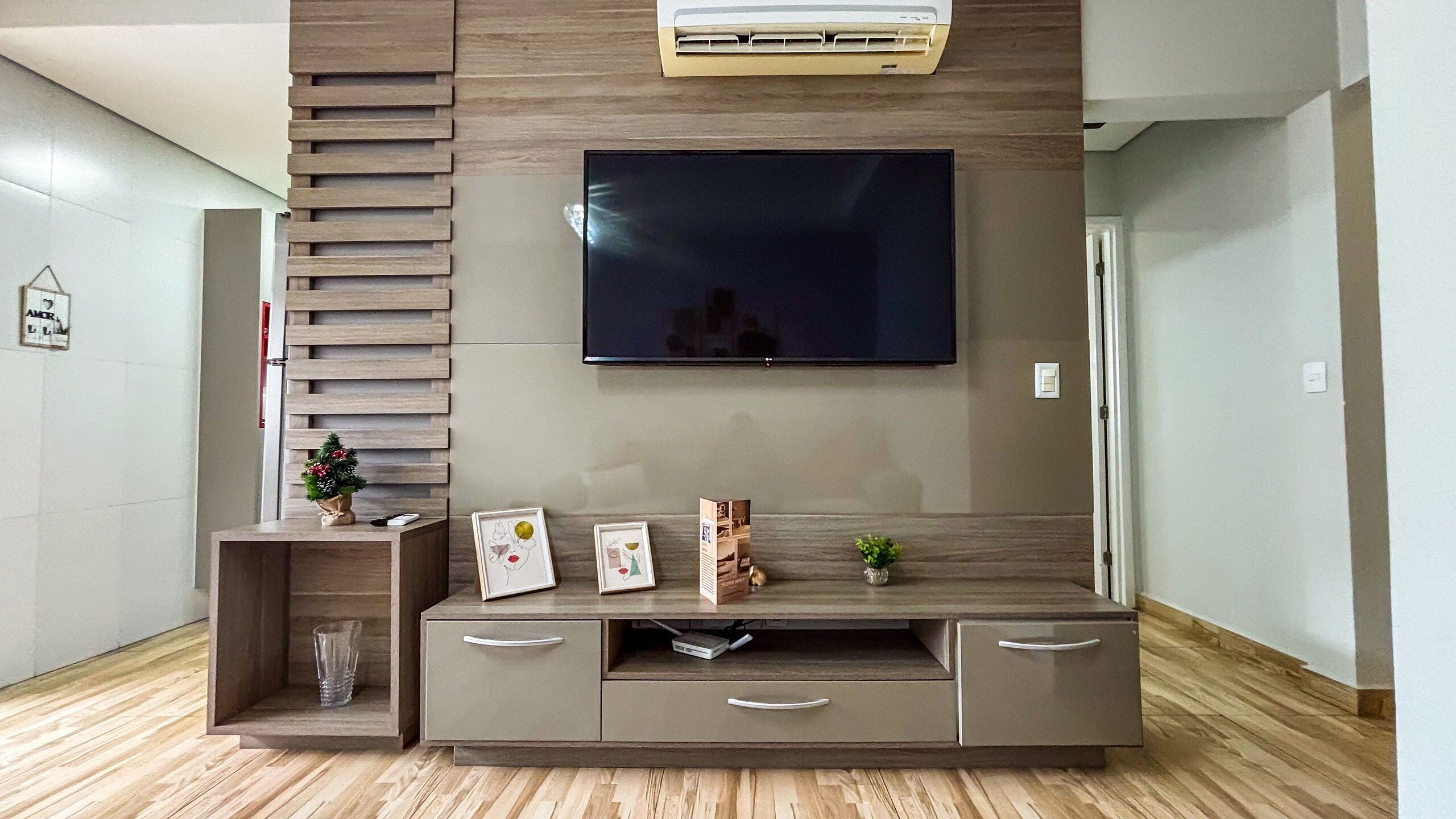 A modern living room with a flat-screen TV mounted on a wooden wall, a built-in entertainment unit with drawers, and a small plant on the side.