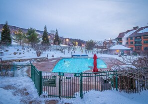 Ein Swimmingpool, umgeben von verschneierter Landschaft, mit einem Zaun, einem roten Sonnenschirm und einem Gebäude im Hintergrund.
