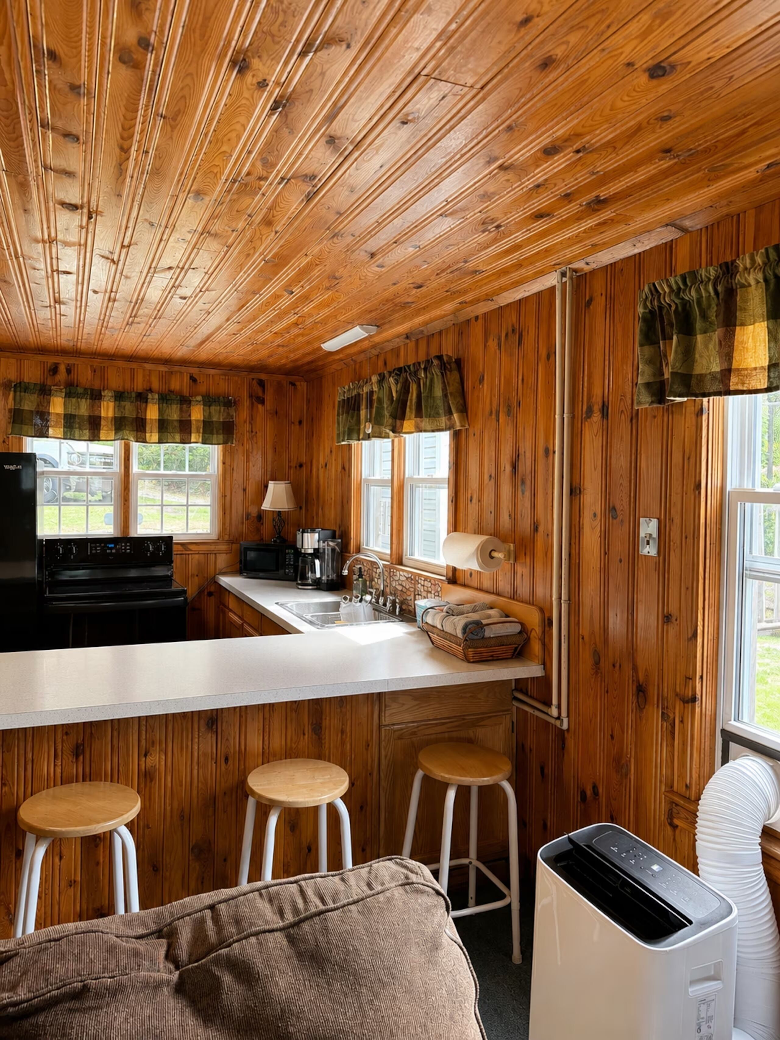 Interior de una cabaña con zona de cocina, paredes de madera, un taburete y una ventana con cortinas.
