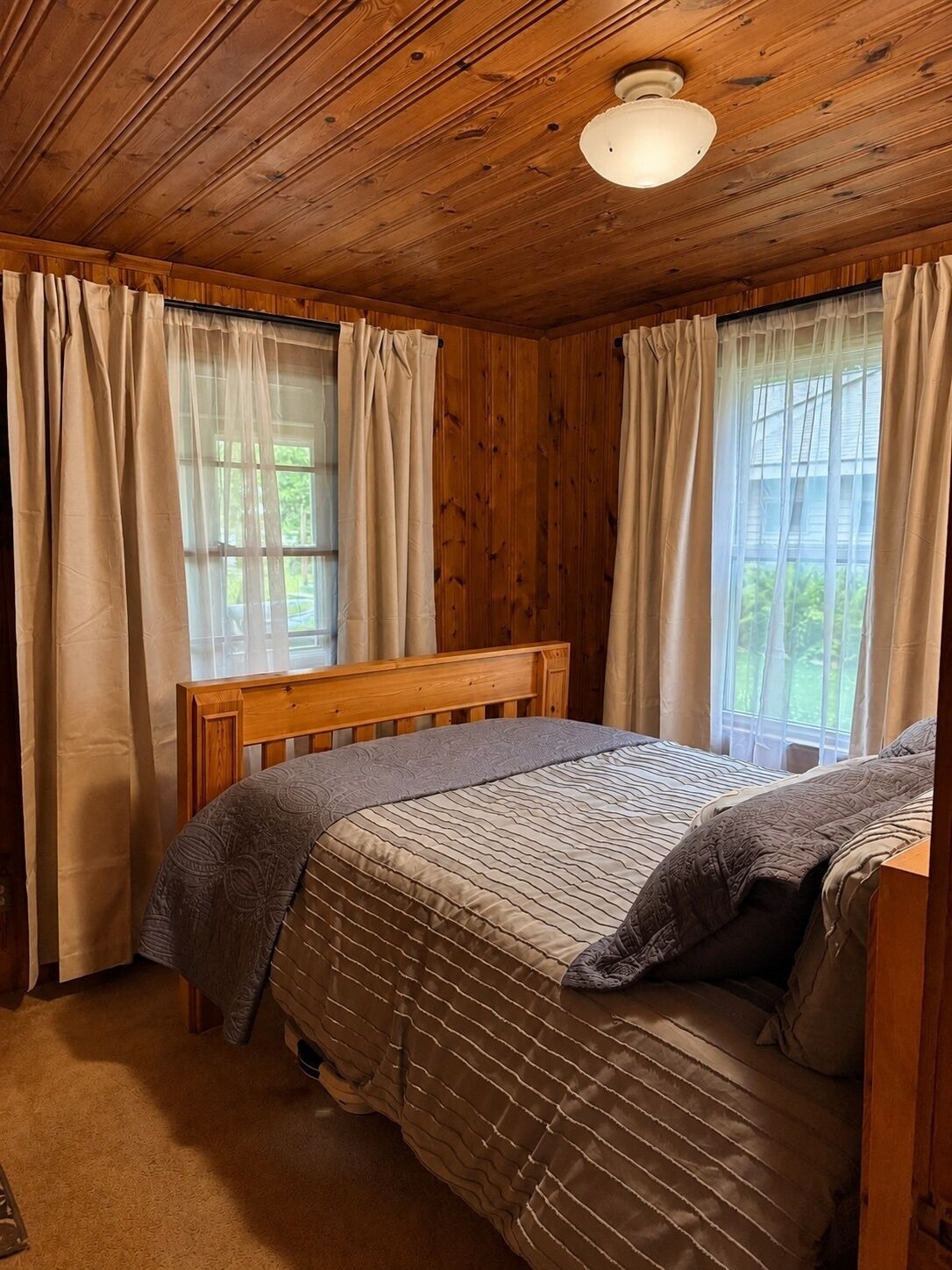 A wooden-paneled room with a bed, a window with curtains, and a ceiling light.