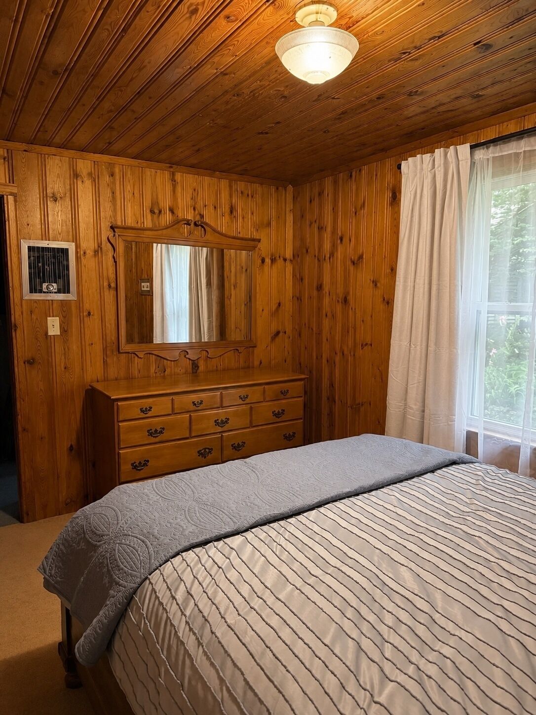 A wooden-paneled room with a bed, dresser, mirror, and window with curtains.