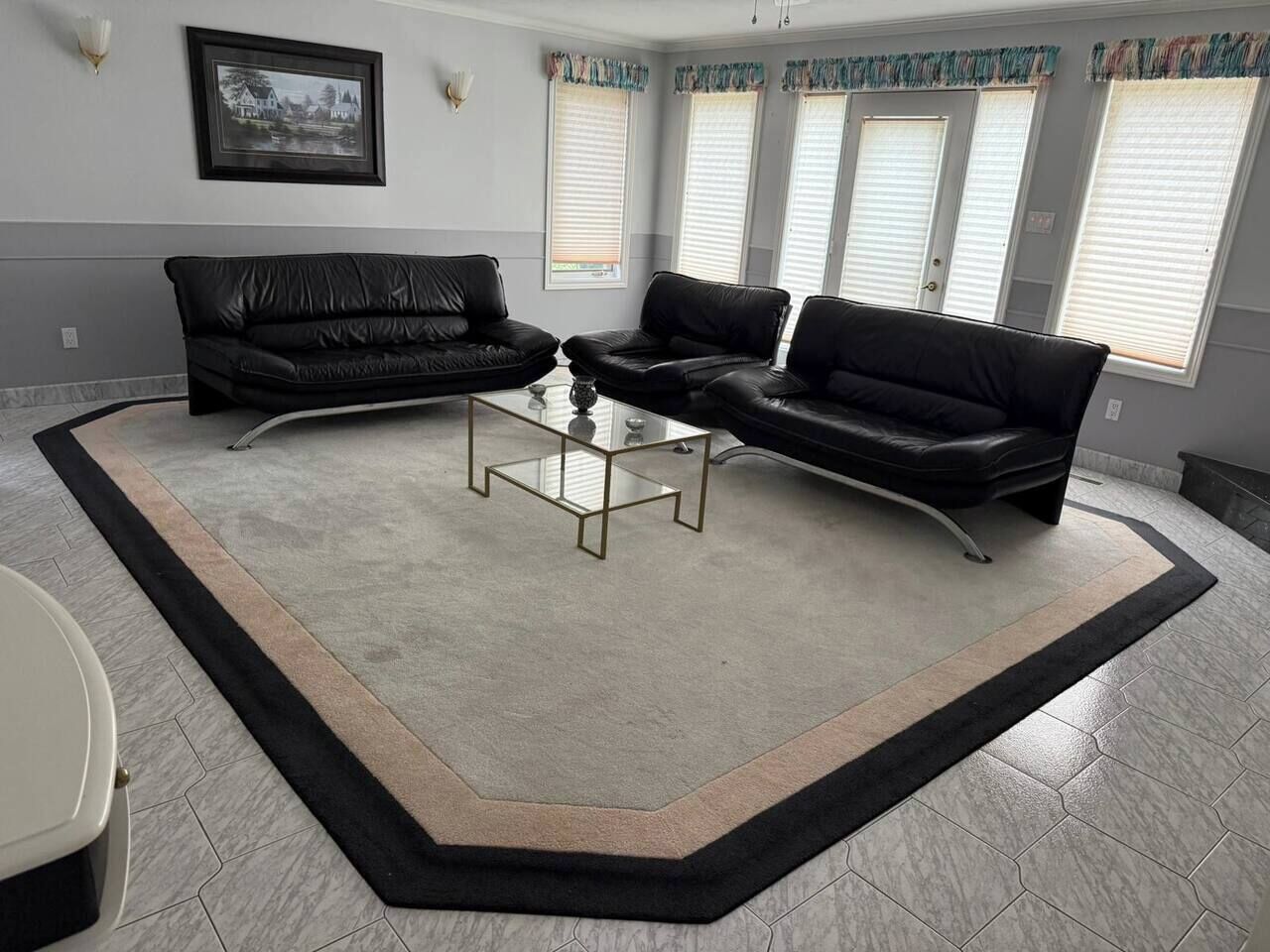 Sala de estar com sofá de couro preto, mesa de centro de vidro e uma grande janela com persianas.