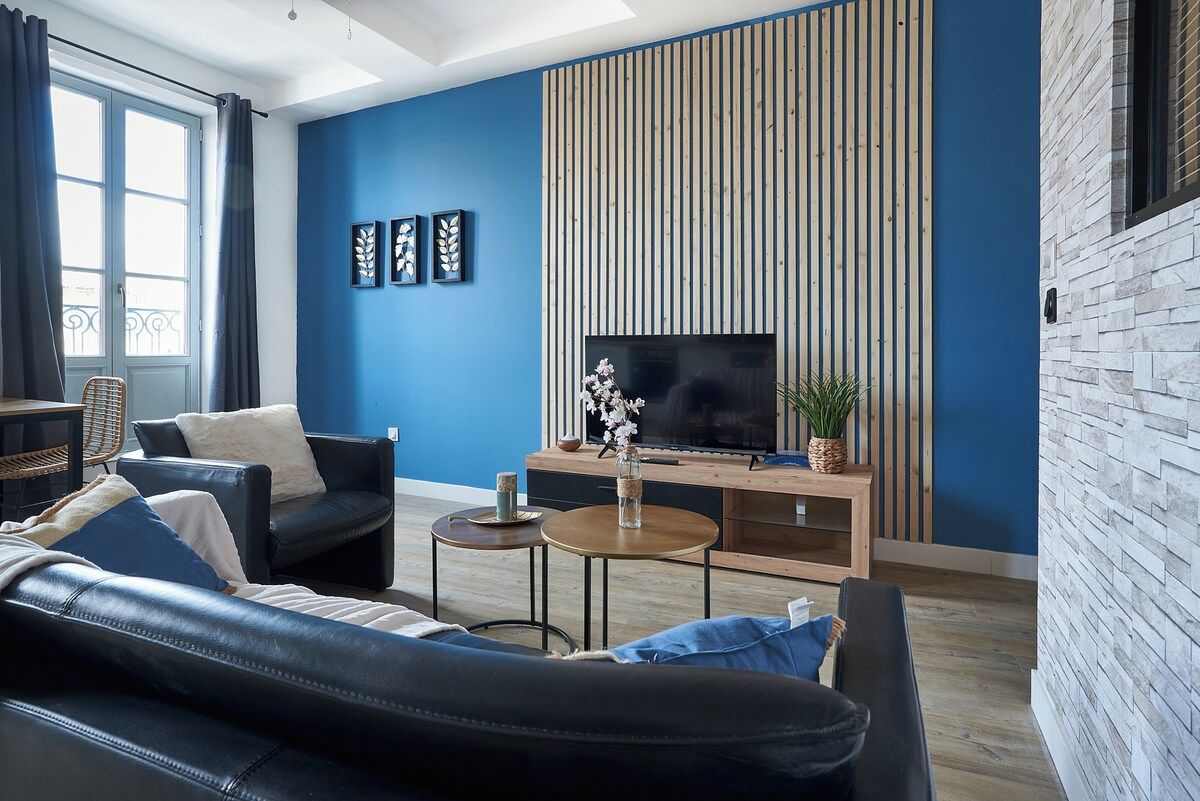 A modern living room with a blue wall, a wooden slat wall, a black leather sofa, a TV, and a small table with a plant.