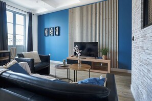 A modern living room with a blue wall, a wooden slat wall, a black leather sofa, a TV, and a small table with a plant.