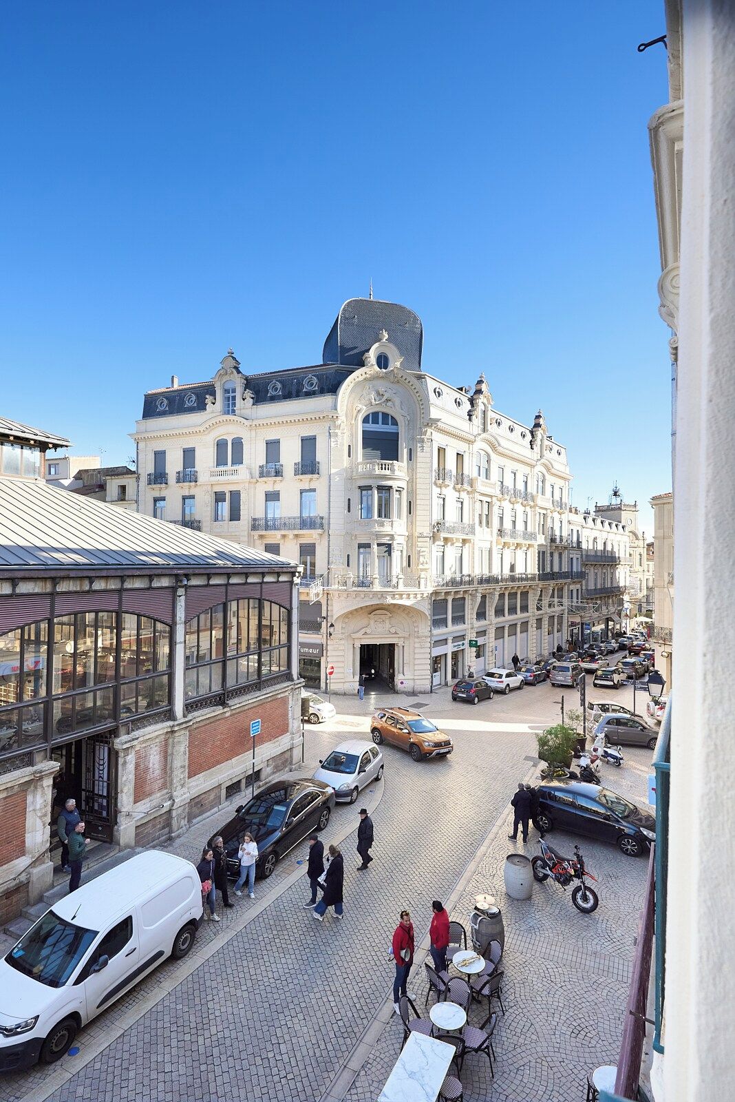 A historic building with a central entrance, a covered area with tables and chairs, and a street with cars and pedestrians.