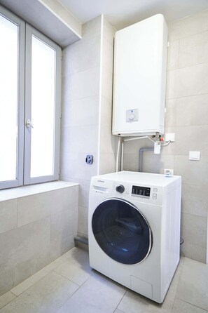 A compact laundry room with a front-loading washing machine, a built-in heater, and a window. - Cozy stay place de la Madeleine (Béziers)